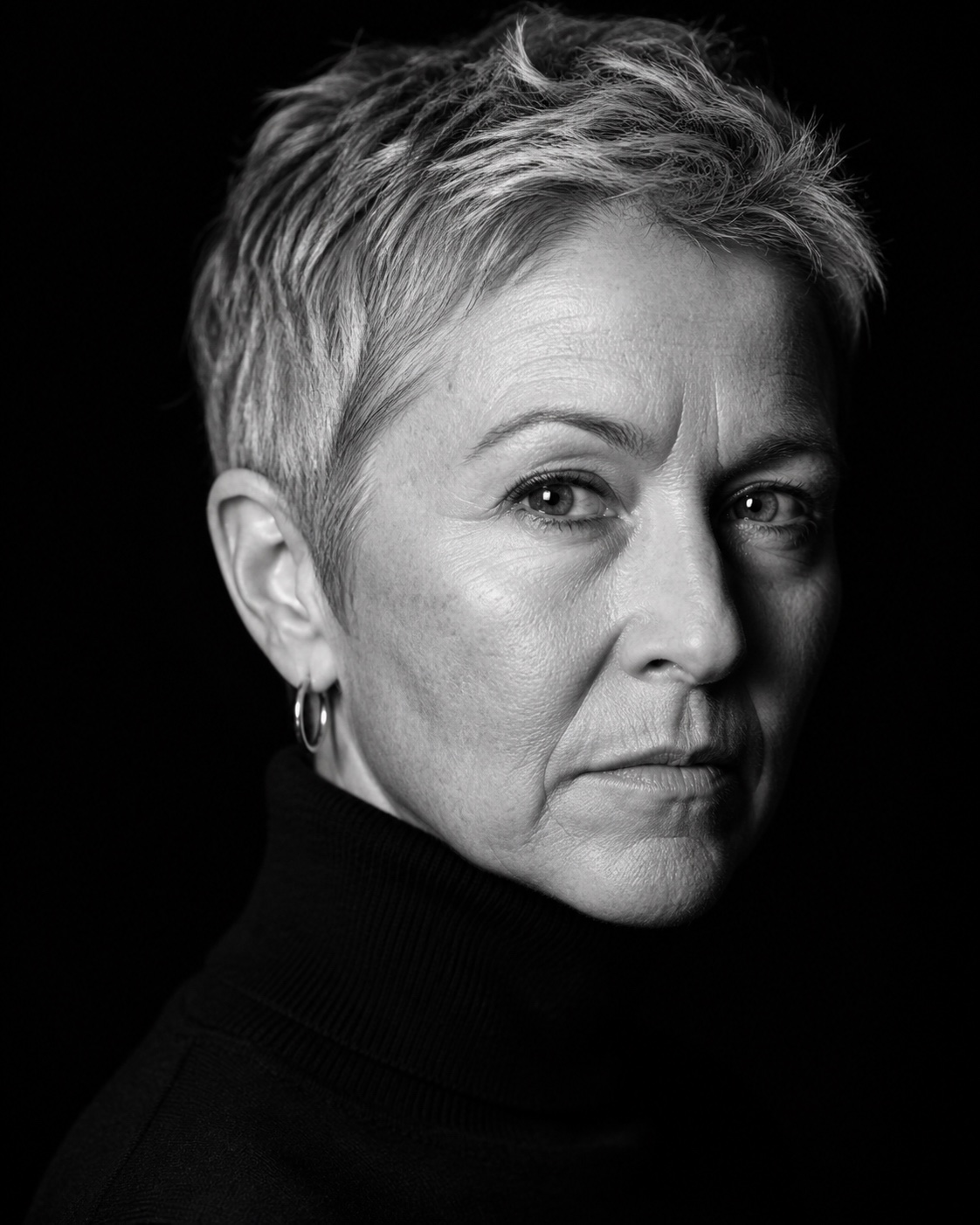 High-contrast black and white studio portrait of a woman with cropped silver hair in a black turtleneck under Rembrandt lighting